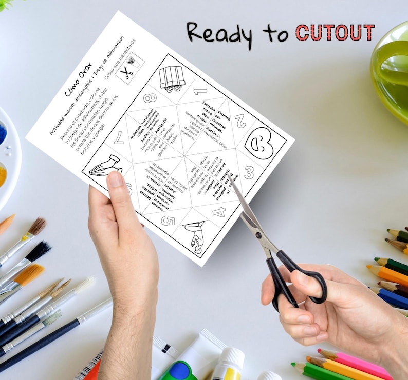 May include: A white paper with the text "C&oacute;mo Orar" and "Ready to CUTOUT" is being cut with scissors. The paper has a geometric design with numbers and illustrations. Paintbrushes and colored pencils are in the background.