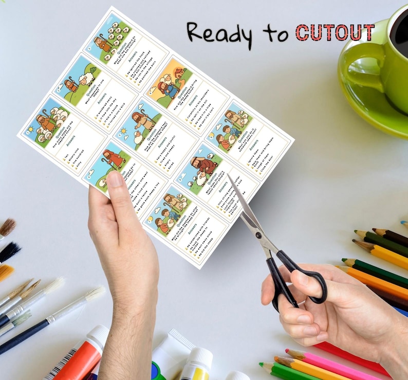 May include: A sheet of paper with illustrated cards featuring biblical scenes and text, held by a person's hands. The words "Ready to CUTOUT" are written at the top. Scissors are cutting the paper. Colored pencils, paint tubes, and a green mug are in the background.