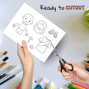 May include: A white paper with black line drawings of religious figures and symbols, including a person, sandals, a cross, and a robe. The text "Ready to CUTOUT" is written at the top. Scissors are cutting the paper.