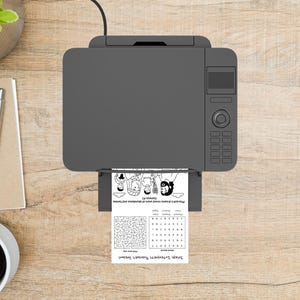 May include: A gray printer is printing a black and white page with illustrations and text. The page includes a maze and the text "Joseph Interprets Pharaoh's Dreams". The printer is on a light brown wooden surface.