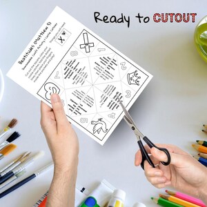 May include: A white paper craft activity sheet with the text "Beatitudes (Matthew 5)" and "Ready to CUTOUT". The sheet is being cut with black-handled scissors. Paintbrushes, paint tubes, and colored pencils are in the background.
