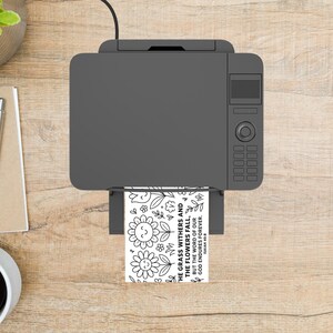May include: A gray printer is printing a black and white coloring page with floral designs and the text "THE GRASS WITHERS AND THE FLOWERS FALL, BUT THE WORD OF OUR GOD ENDURES FOREVER. ISAIAH 40:8". The printer sits on a light wood surface.