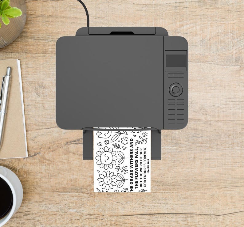 May include: A gray printer is printing a black and white coloring page with floral designs and the text "THE GRASS WITHERS AND THE FLOWERS FALL, BUT THE WORD OF OUR GOD ENDURES FOREVER. ISAIAH 40:8". The printer sits on a light wood surface.