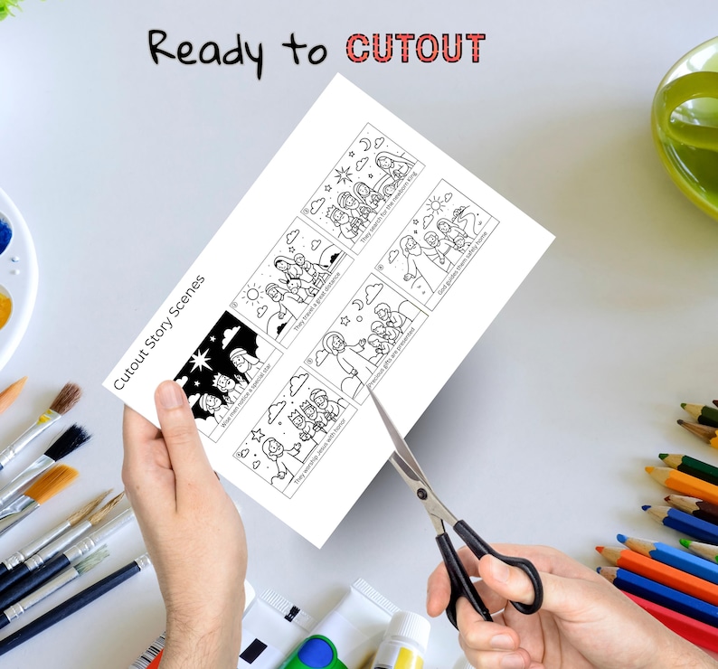 May include: A white sheet of paper titled Cutout Story Scenes featuring six black and white line art illustrations depicting the nativity story with text captions. The paper is being cut with black metal scissors, surrounded by various art supplies.