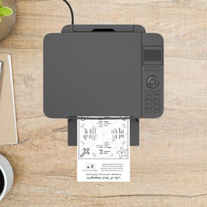 Op de afbeelding: Een grijze printer die een zwart-wit vel papier afdrukt met de tekst "Life of Jesus Disciples" en een waarzeggerontwerp. De printer staat op een houten oppervlak, met een pen, notitieblok en koffiekopje zichtbaar.