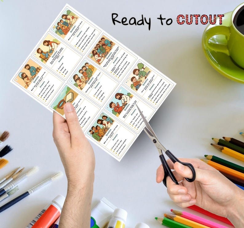 May include: A sheet of illustrated cards with religious themes is being cut with scissors. The cards feature colorful drawings and text. The words "Ready to CUTOUT" are written in red and black. A green mug and colored pencils are in the background.