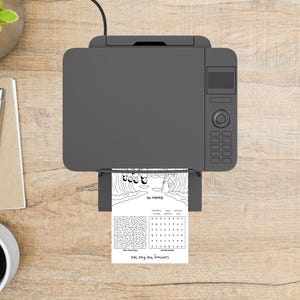 May include: A top-down view of a gray printer printing a black and white activity sheet. The sheet features a maze, word search, and an illustration of people crossing the Red Sea. The printer is on a wooden surface.