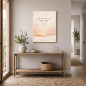 Può includere: Una stampa incorniciata con il testo "HIS MERCIES ARE NEW" è appesa sopra una consolle in legno. La stampa presenta un paesaggio in stile acquerello nei toni del pesca e del crema. Un cesto intrecciato e un vaso di ceramica con verde sono sul tavolo.