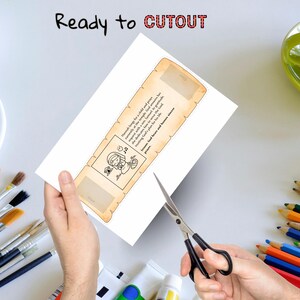 May include: A white paper bookmark with a scroll design, featuring text and a cartoon illustration. The words "Ready to CUTOUT" are at the top. Scissors are cutting the bookmark. Paintbrushes and pencils are in the background.
