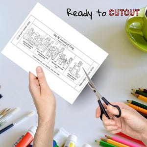 May include: A black and white illustrated paper with the text "David Anointed King" and "Ready to CUTOUT". The paper is being cut with scissors. A green mug and colored pencils are in the background.