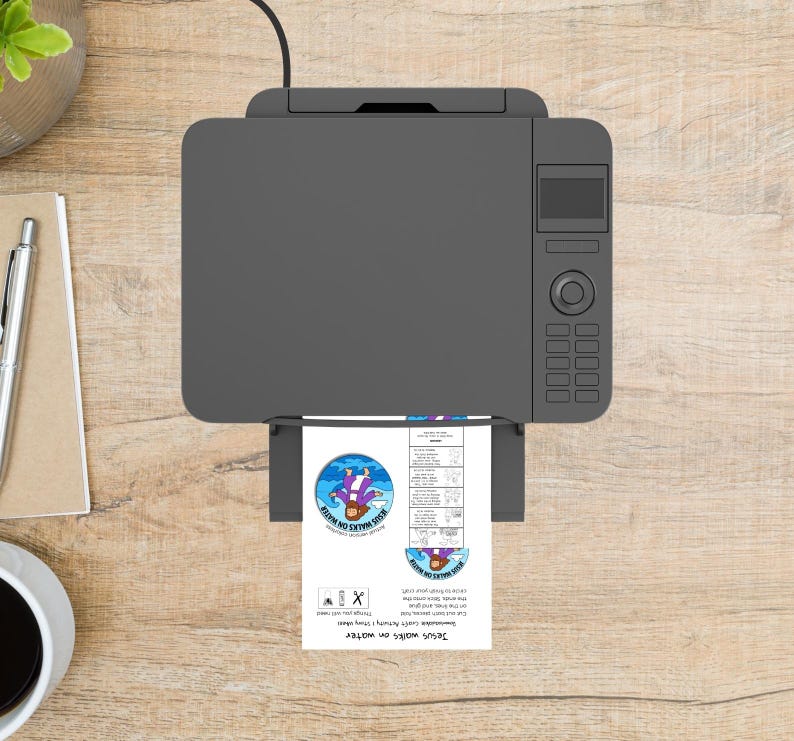 May include: A gray printer with a printed sheet of paper. The paper has a blue and purple graphic with the text "Jesus walks on water". The printer is on a light brown wooden surface.