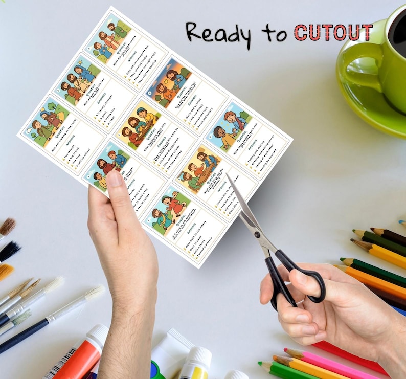 May include: A sheet of illustrated cards with questions and answers, held by a person's hands, with scissors cutting the cards. The text "Ready to CUTOUT" is printed at the top. A green mug and art supplies are in the background.