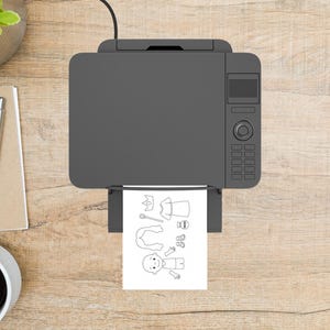 May include: A gray printer is printing a black and white coloring page of a cartoon character and accessories. The printer is on a light brown wooden surface. A pen, notepad, and coffee cup are visible.