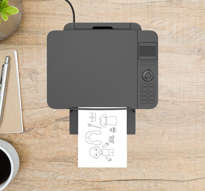 May include: A gray printer is printing a black and white coloring page of a cartoon character and accessories. The printer is on a light brown wooden surface. A pen, notepad, and coffee cup are visible.