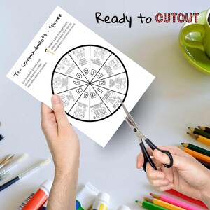 Puede incluir: Un papel blanco con un dise&ntilde;o de ruleta de los Diez Mandamientos se est&aacute; cortando con tijeras. La ruleta presenta secciones numeradas con ilustraciones y el texto "Ten Commandments - Spinner" y "Ready to CUTOUT".