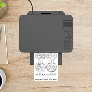 May include: A gray printer is printing a sheet with four circular illustrations and text. The text reads "Parable of the Workers in the Vineyard Story Ornament". The printer is on a light brown wooden surface.