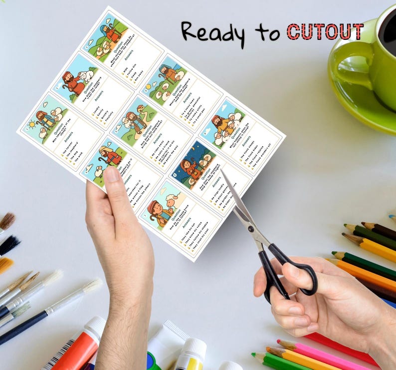 May include: A sheet of paper with illustrated cards featuring biblical scenes, held by hands with scissors cutting the cards. The text "Ready to CUTOUT" is printed at the top. A green mug and coloured pencils are in the background.