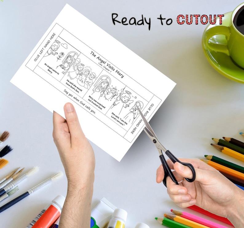 May include: A black and white illustrated cutout sheet titled "The Angel Visits Mary" with text and images. The sheet is being cut with scissors. The words "Ready to CUTOUT" are written at the top. A green mug of coffee and colored pencils are in the background.