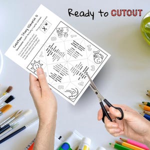 May include: A black and white printable "Creation Story" cootie catcher with the text "Ready to CUTOUT". The cootie catcher is being cut with scissors. Paintbrushes, paint tubes, and colored pencils are in the background.