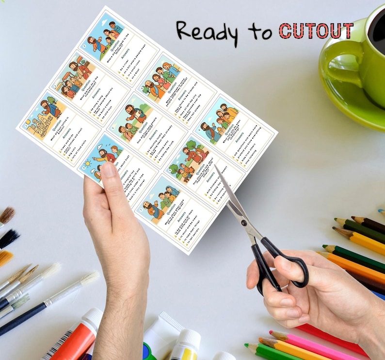 K&ouml;nnte beinhalten: Ein Bogen mit bedruckten Karten mit Illustrationen und Text wird mit einer Schere geschnitten. Die Karten zeigen farbenfrohe Zeichnungen von Menschen und Text. Die Worte "Ready to Cutout" sind oben aufgedruckt. Farbtuben, Pinsel und Buntstifte sind im Hintergrund.