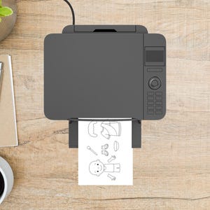 May include: A grey printer, viewed from above, is printing a black and white colouring page featuring a baby and clothing items. The printer sits on a light brown wooden surface, with a pen, notepad, and coffee cup visible.