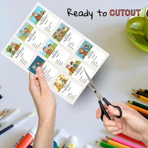 May include: A sheet of printed cards with illustrations and text, held by a person's hand, being cut with scissors. The cards feature images of people and text that reads "Ready to CUTOUT". A green mug and colored pencils are in the background.