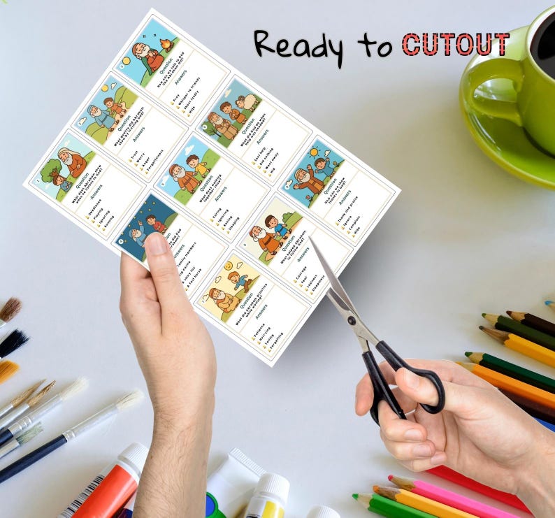 May include: A sheet of printed cards with illustrations and text, held by a person's hand, being cut with scissors. The cards feature images of people and text that reads "Ready to CUTOUT". A green mug and colored pencils are in the background.