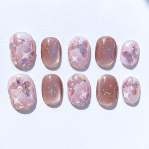 May include: A collection of ten artificial nails. Some are a solid, shimmering brown colour, while others have a pink base with glitter and star-shaped details. The nails are oval and arranged on a white surface.