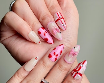May include: A set of almond-shaped artificial nails with various designs. Some nails are solid beige, others feature red hearts on a pink background, and some have pink bows and red plaid patterns. Rhinestones add sparkle.