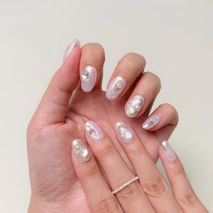 Op de afbeelding: Close-up van een hand met lange, ovaalvormige nagels. De nagels zijn gelakt met een parelmoer witte lak en versierd met iriserende edelstenen, parels en zilveren accenten. Een zilveren ring met kleine steentjes zit aan de ringvinger.