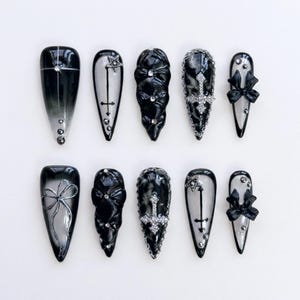 May include: A set of ten black and silver press-on nails. Designs include a cross, star, bow, and gradient effect. Embellishments include rhinestones and chains. The nails are almond-shaped.
