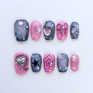 May include: A set of ten artificial nails in various designs. The nails feature a mix of pink and gray colors, adorned with embellishments like stars, hearts, bows, and pearls. Some nails have a glossy finish, while others have a matte appearance.