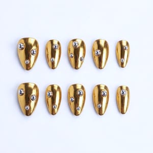 May include: Ten gold-colored, almond-shaped press-on nails with silver-colored, spherical embellishments. The nails are arranged in two rows of five, showcasing a glossy finish. The design features a metallic, polished look.