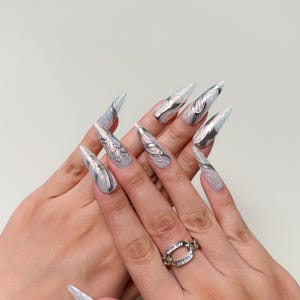 May include: Long, pointed artificial nails with a silver and white design. The nails have a metallic silver accent and a swirling pattern. A silver ring with clear stones is on the ring finger.