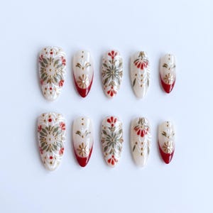 Può includere: Set di dieci unghie finte con base color crema, decorate con motivi floreali dorati, rossi e verdi. Ogni unghia ha una forma arrotondata e una punta rossa. Le unghie sono disposte su uno sfondo bianco.