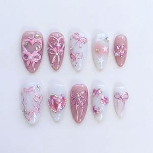 May include: A set of ten artificial nails in various designs. The nails feature a combination of pink and white colors, adorned with bows, hearts, stars, and rhinestones. The nail shapes are oval and almond, with a glossy finish.
