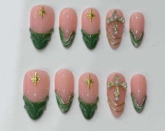 May include: Set of ten artificial nails in a variety of designs. The nails feature a pink base with gold accents, green leaf-like designs, and gold star or cross embellishments. The nails are arranged on a white surface.