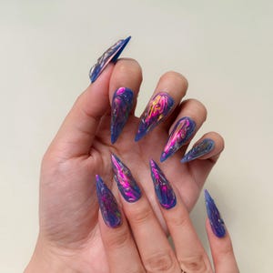 May include: A hand showing long, pointed, iridescent acrylic nails. The nails are a deep blue with swirling pink, purple, and yellow highlights, creating a holographic effect. The nails are a stiletto shape.
