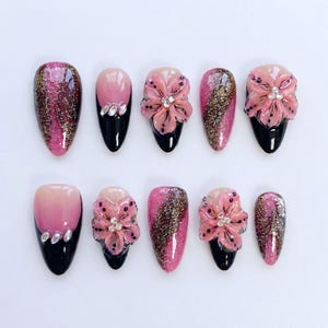 Peut inclure: Un ensemble de dix faux ongles aux designs variés. Certains ongles présentent un motif pailleté rose et or, d'autres ont un effet dégradé rose, et certains sont ornés d'accents de fleurs roses en 3D et de strass. Les bouts des ongles sont peints en noir.