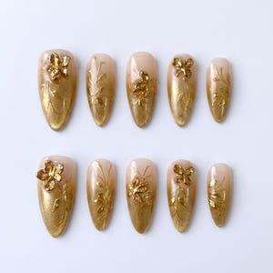 May include: Set of ten artificial nails with a gold gradient design. Each nail features gold floral embellishments and intricate leaf patterns. The nails have a glossy finish and are arranged on a white surface.