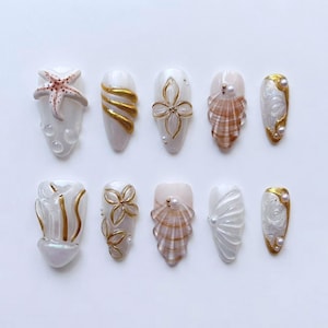 May include: A collection of ten artificial nails with a seaside theme. Designs include a starfish, gold swirls, flower and shell accents, and pearl embellishments. The nails are white, gold, and brown, with a glossy finish.