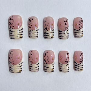 May include: A set of ten artificial nails. Each nail features a light pink top with black speckles and small pink flower accents. The lower half has a cream-colored tiger stripe design with gold accents.