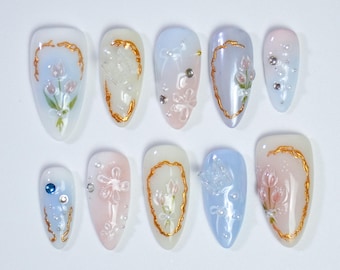 Op de afbeelding: Een set van tien kunstnagels in verschillende vormen en maten. Elke nagel heeft een ander ontwerp, waaronder bloempatronen, gouden accenten en iriserende versieringen. Het kleurenpalet omvat zachte pasteltinten zoals roze, blauw en wit.