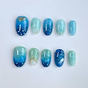 Puede incluir: Un conjunto de diez uñas artificiales con un diseño azul y verde inspirado en el océano. Cada uña presenta un acabado brillante con diferentes patrones, incluyendo puntos blancos y acentos de lámina dorada. Las uñas son de forma ovalada.
