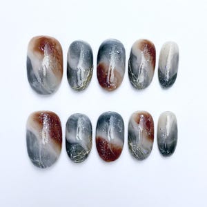 Peut inclure: Un ensemble de dix faux ongles ovales. Chaque ongle présente un motif marbré avec des nuances de marron, de gris et de blanc, agrémenté de paillettes argentées et de veinures dorées. Les ongles sont disposés sur une surface blanche.