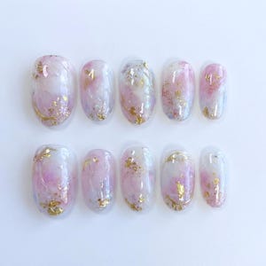 May include: A set of ten artificial nails with a glossy finish. Each nail features a blend of pink, white, and gold glitter accents. The nails have an oval shape and are arranged on a white surface.