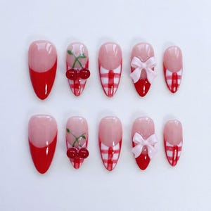 May include: A set of ten artificial nails in various designs. Some nails feature a red French tip, while others showcase cherry and bow accents. The nails have a glossy finish and are arranged on a white surface.