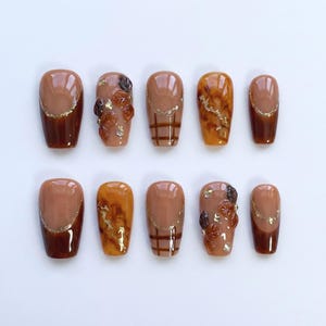 May include: A set of ten artificial nails in various designs. The nails feature a mix of brown, tan, and gold colors. Some nails have gold glitter accents, while others have leaf and plaid patterns. The nails are oval-shaped.