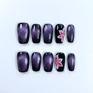 May include: A set of ten press-on nails. The nails are a deep purple with a cat-eye effect, and two nails feature a black base with a pink floral design and rhinestone accents. The nails are oval-shaped.