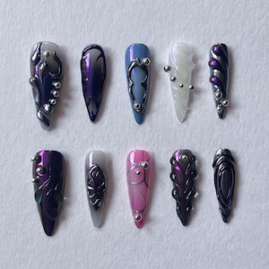Könnte beinhalten: Eine Sammlung von zehn künstlichen Nägeln in verschiedenen Designs und Farben. Die Nägel weisen eine Reihe von Farben auf, darunter Lila, Blau, Rosa und Silber, mit dekorativen Elementen wie metallischen Akzenten und Wirbelmustern.